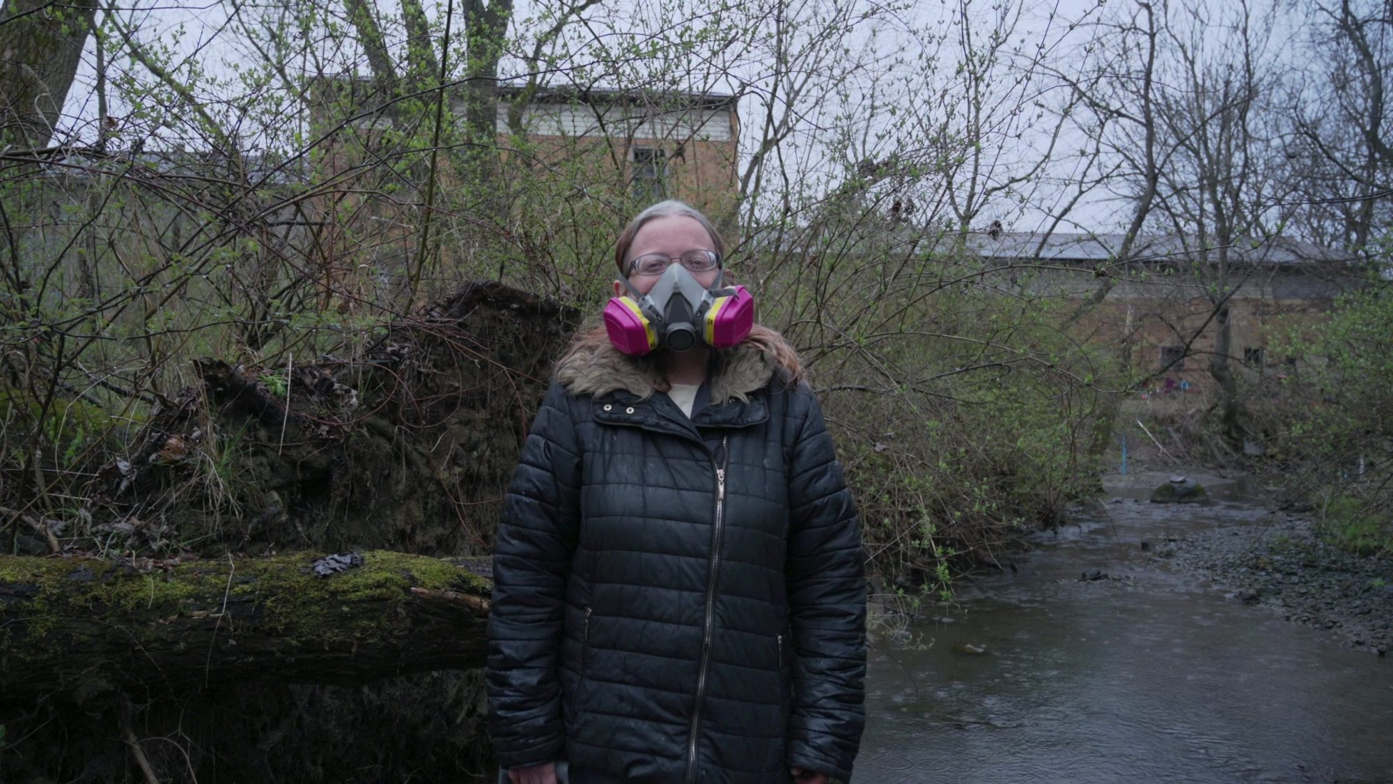 Still image from TRNN's documentary report "Trainwreck in 'Trump Country,'" featuring Christina Siceloff, a resident of Beaver County, PA, standing in a creek in East Palestine, OH, with a pink gas mask on her face. Image courtesy of Mike Balonek.