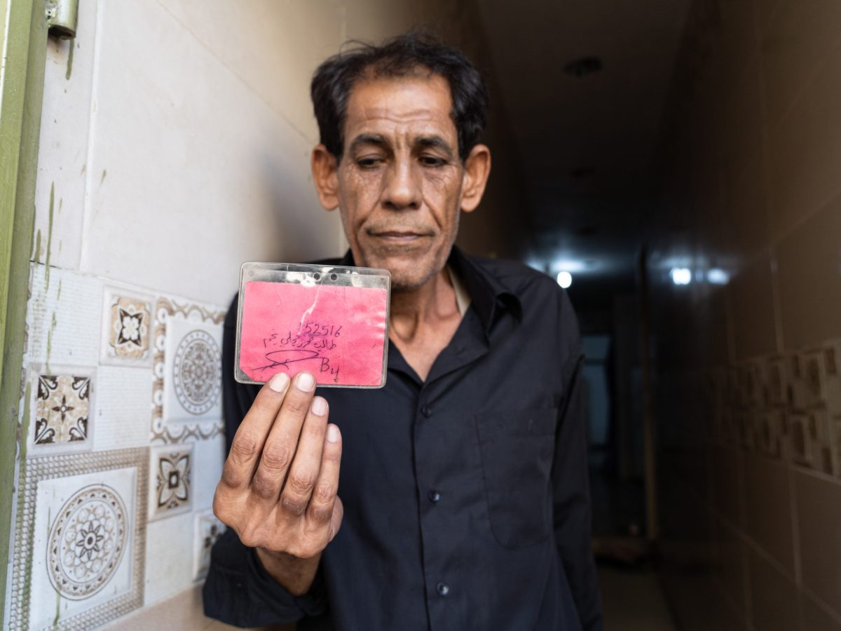 Taleb al-Majli holding a prisoner identity card that he says US forces issued to him at Abu Ghraib. One side lists his inmate number, name, and cell number. The other side shows an official prison stamp received upon entering Abu Ghraib.