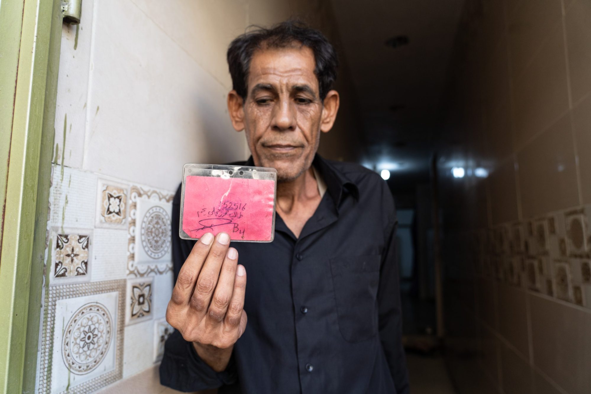 Taleb al-Majli holding a prisoner identity card that he says US forces issued to him at Abu Ghraib. One side lists his inmate number, name, and cell number. The other side shows an official prison stamp received upon entering Abu Ghraib.