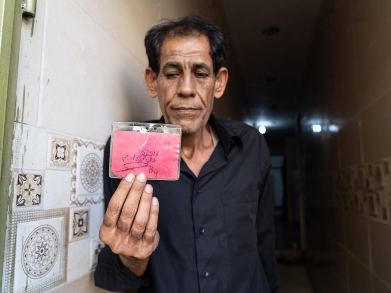 Taleb al-Majli holding a prisoner identity card that he says US forces issued to him at Abu Ghraib. One side lists his inmate number, name, and cell number. The other side shows an official prison stamp received upon entering Abu Ghraib.
